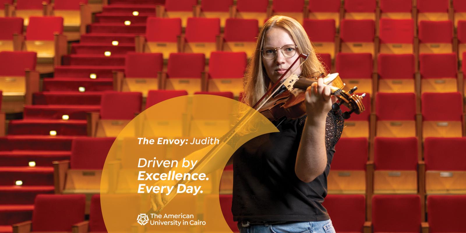 A female wearing glasses playing the violin in front of stage seats. Text reads "The Envoy: Judith. Driven by Excellence. Every Day. ϲͼ"