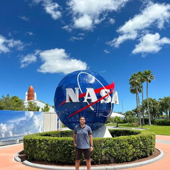 A male is smiling and standing in front of the NASA globe, there are palm trees and a rocket behind him