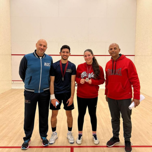 Three males and a female standing in a squash court, one of the males and the female are wearing medals